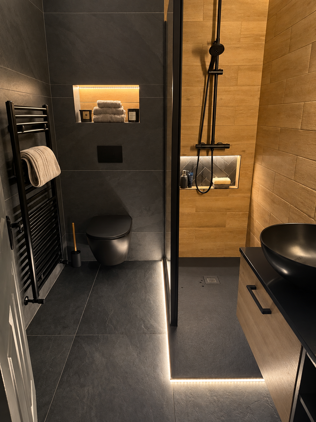 Modern dark bathroom with black fittings, heated towel rail, glass shower, wood accent wall and black vessel basin
