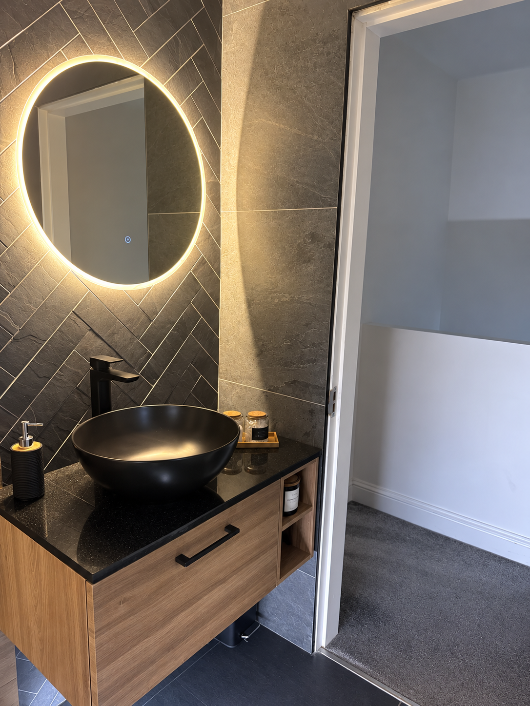 Black vessel basin and tap on a timber vanity with dark herringbone tiles and an illuminated round mirror